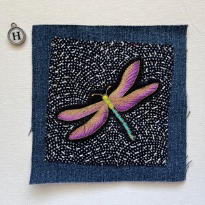 May include: A denim patch with a dragonfly design. The dragonfly has purple wings with gold accents, a yellow head, and a turquoise body. The background is black with white dots. A silver charm with the letter "H" is in the upper left corner.