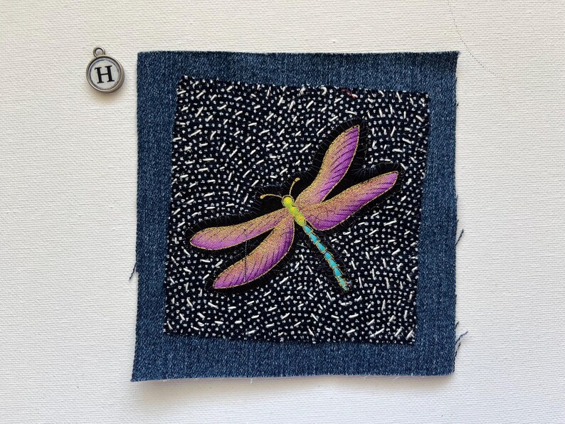 May include: A denim patch with a dragonfly design. The dragonfly has purple wings with gold accents, a yellow head, and a turquoise body. The background is black with white dots. A silver charm with the letter "H" is in the upper left corner.