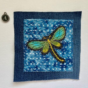 May include: A square denim patch with a hand-stitched dragonfly design. The dragonfly has teal and green wings, a yellow body, and black outlines. The background is blue with white cross-stitch details. A small metal charm with the letter "A" is in the upper left corner.