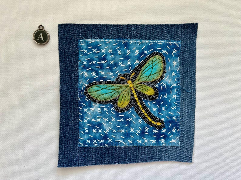 May include: A square denim patch with a hand-stitched dragonfly design. The dragonfly has teal and green wings, a yellow body, and black outlines. The background is blue with white cross-stitch details. A small metal charm with the letter "A" is in the upper left corner.