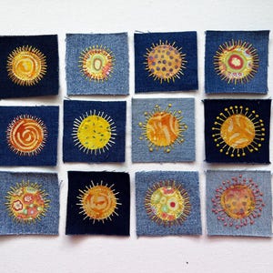 May include: Twelve square denim fabric patches arranged in a grid. Each patch features a circular sun motif created with colorful fabric appliqués and yellow embroidered rays radiating outward. The denim backgrounds vary in shades of blue.