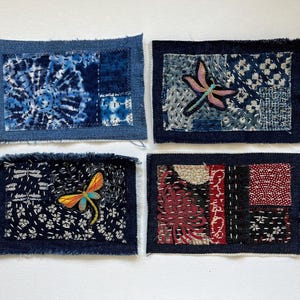 May include: Four small, rectangular textile art pieces with frayed edges. Each features a dark blue denim border and a central design. One has a tie-dye pattern, two have dragonfly embroidery, and one has a floral pattern.