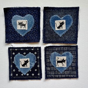 May include: Four square, dark blue fabric patches with a heart-shaped denim appliqué. Each heart features a black cat illustration on a white background. The patches have different patterned backgrounds and are hand-stitched.