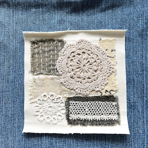 May include: A square fabric patch with various textured elements. It features a large, beige crocheted doily, smaller patches of textured fabric, and white and gray lace. The patch is set against a blue denim background.