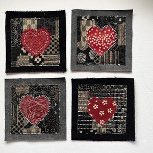 May include: Four square fabric patches with a heart design. Each patch has a different pattern and a red heart in the center. The hearts are stitched onto the patches. The patches are framed with a dark fabric.