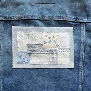May include: Close-up of a denim jacket back with a patchwork design. The patch features various fabric squares in shades of blue, white, and cream, with floral and geometric patterns. The jacket has visible stitching and a classic denim texture.