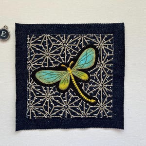 May include: A square denim patch with a dragonfly applique. The dragonfly has turquoise wings and a yellow body, set against a black background with a geometric pattern. A small silver charm with the letter "E" is in the upper left corner.