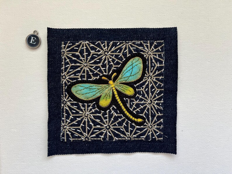 May include: A square denim patch with a dragonfly applique. The dragonfly has turquoise wings and a yellow body, set against a black background with a geometric pattern. A small silver charm with the letter "E" is in the upper left corner.