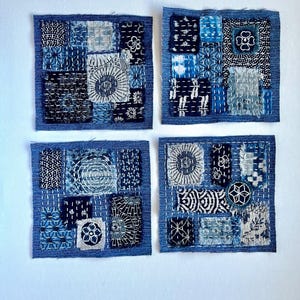 May include: Four square textile art pieces with a denim blue border. Each square features a patchwork design with various shades of blue and white fabrics, showcasing intricate stitching and patterns. The overall aesthetic is handcrafted and artistic.