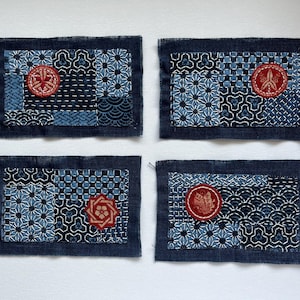 May include: Four rectangular fabric patches with intricate blue and white geometric patterns and red circular embellishments. The patches are framed by a dark blue border, showcasing a blend of textures and designs.