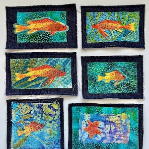 May include: Six fabric art pieces featuring colorful fish designs. Each piece has a dark blue denim border and a vibrant, patterned background in shades of blue and green. The fish are primarily orange and yellow, with intricate details.