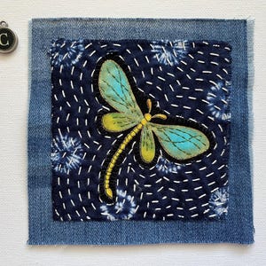 May include: A denim patch with a dragonfly design. The dragonfly has teal and yellow wings, a yellow body, and black outlines. The patch is stitched onto a dark blue fabric with white stitching and a denim border. A silver charm with the letter "C" is in the upper left corner.