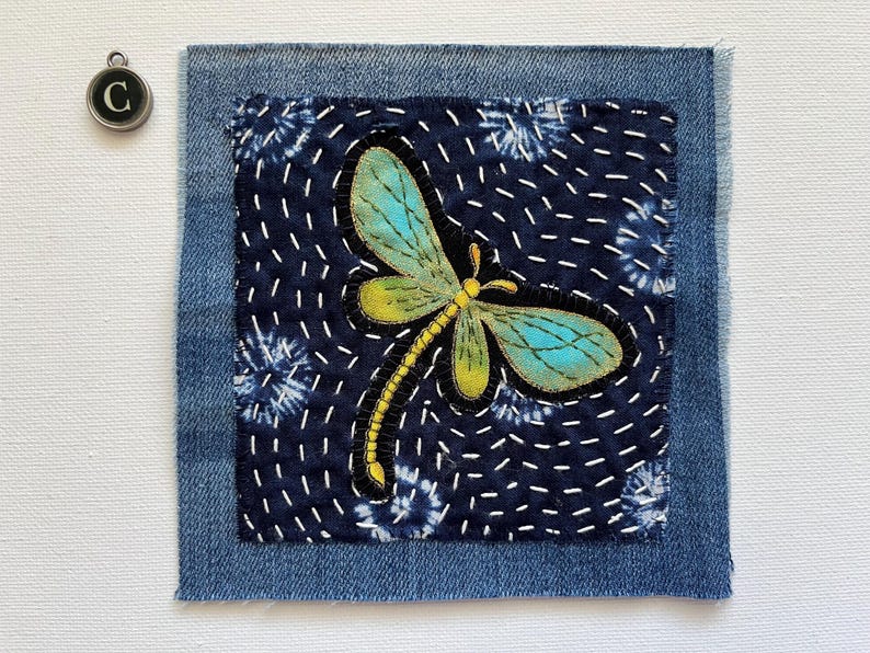 May include: A denim patch with a dragonfly design. The dragonfly has teal and yellow wings, a yellow body, and black outlines. The patch is stitched onto a dark blue fabric with white stitching and a denim border. A silver charm with the letter "C" is in the upper left corner.