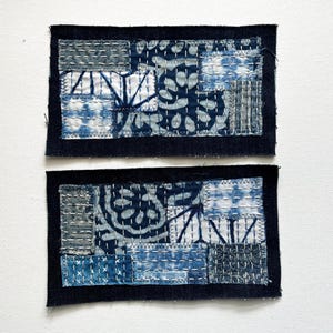 May include: Two rectangular patches with a dark blue border. The patches are composed of smaller fabric pieces in shades of blue and white, stitched together with white thread. The design features geometric and floral patterns, creating a patchwork aesthetic.