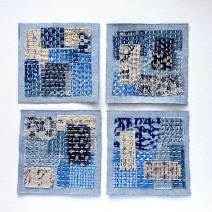 May include: Four square fabric coasters featuring a patchwork design of various blue and cream textile scraps. Each coaster is constructed with visible white running stitch embroidery, creating a textured surface with diverse geometric and floral patterns.