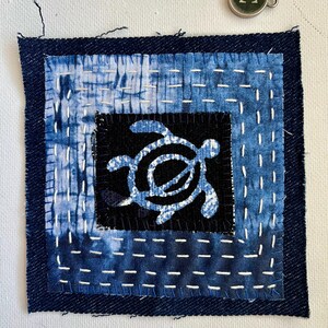 Peut inclure: Une pi&egrave;ce d'art textile carr&eacute;e avec un motif de tortue. L'&oelig;uvre utilise du tissu teint &agrave; l'indigo avec des coutures blanches visibles. La tortue est dans un carr&eacute; noir, entour&eacute;e de tissu &agrave; motifs bleu et blanc, et d'une bordure bleu fonc&eacute;. Un petit charme en m&eacute;tal est dans le coin sup&eacute;rieur droit.