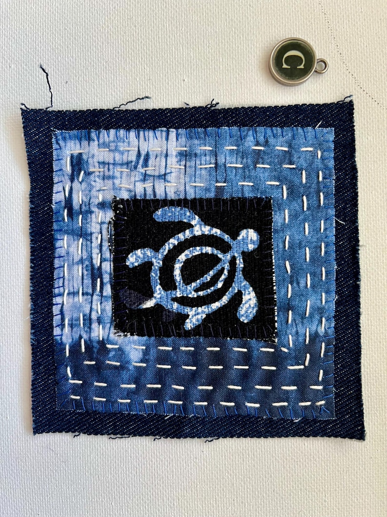 Peut inclure: Une pi&egrave;ce d'art textile carr&eacute;e avec un motif de tortue. L'&oelig;uvre utilise du tissu teint &agrave; l'indigo avec des coutures blanches visibles. La tortue est dans un carr&eacute; noir, entour&eacute;e de tissu &agrave; motifs bleu et blanc, et d'une bordure bleu fonc&eacute;. Un petit charme en m&eacute;tal est dans le coin sup&eacute;rieur droit.
