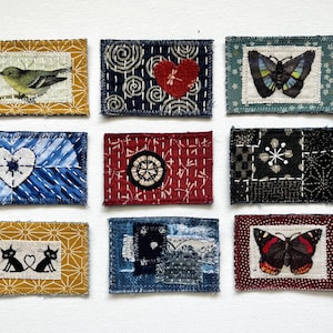 May include: Nine rectangular fabric patches featuring various designs including a bird, a red heart, butterflies, a white heart, a circular emblem, geometric patterns, two black cats with a heart, and layered blue textile scraps with visible stitching.