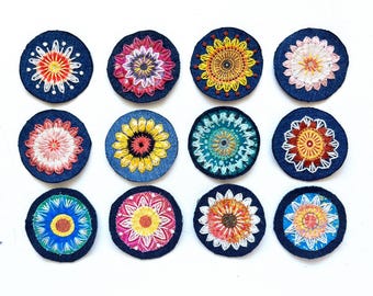 May include: Twelve circular fabric patches featuring intricate, multi-colored floral embroidery on a dark blue denim background. Each patch displays a unique radial pattern with varied thread colors including yellow, red, pink, white, and teal.