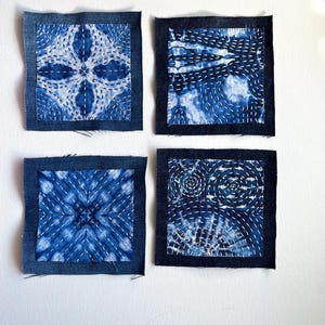 May include: Four square fabric patches with indigo blue and white designs. Each patch has a dark blue denim border and features a unique pattern created with white stitching. The designs include floral, geometric, and spiral motifs.