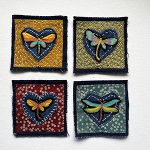 May include: Four square fabric patches with a dragonfly design. Each patch features a denim heart with a dragonfly applique. The backgrounds are mustard yellow, olive green, burgundy floral, and teal floral. The edges are frayed.