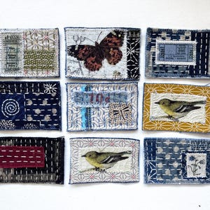 May include: A collection of nine rectangular fabric patches featuring various textile techniques including sashiko stitching, embroidery, and printed motifs such as a butterfly, birds, and floral patterns in shades of blue, yellow, and maroon.