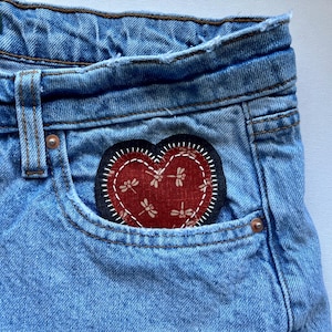 May include: Light blue denim shorts with a heart-shaped patch on the pocket. The patch is red with a dragonfly pattern and white stitching. The shorts have a classic five-pocket design.
