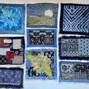 May include: A collection of nine handcrafted textile art pieces. Each square features unique designs, including floral patterns, landscapes, and geometric shapes, all in shades of blue and white. Some pieces have frayed edges and embroidered details, with the word "Dream" on one.