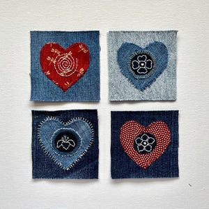 May include: Four denim squares with heart-shaped appliqués. Two hearts are red with white embroidery, and two are blue with black and white floral designs. The squares are arranged in a 2x2 grid.