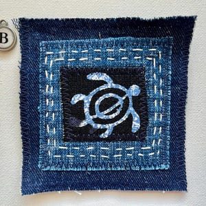 Peut inclure: Un patch carr&eacute; en tissu denim avec un motif de tortue bleu sur fond noir. Le patch est cousu avec du fil blanc et bord&eacute; de bleu fonc&eacute;. Un charm argent&eacute; avec la lettre "B" est &eacute;galement visible.