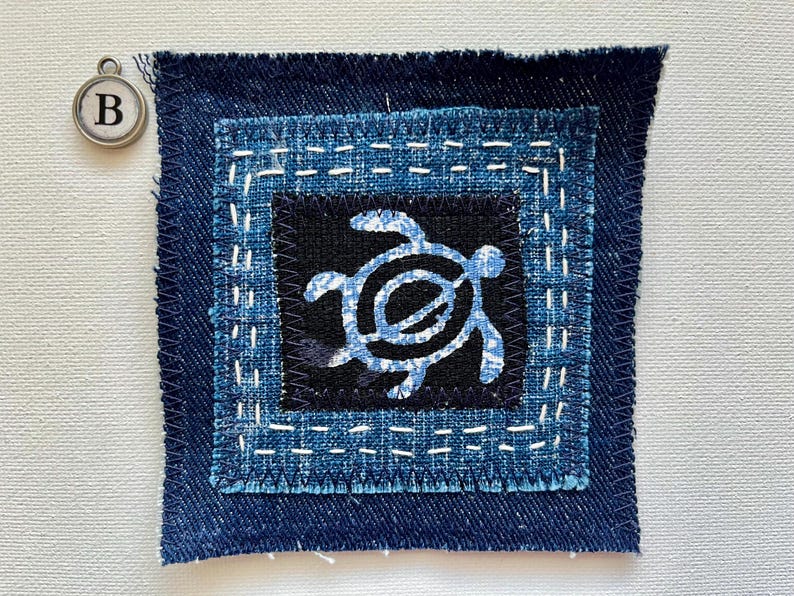 Peut inclure: Un patch carr&eacute; en tissu denim avec un motif de tortue bleu sur fond noir. Le patch est cousu avec du fil blanc et bord&eacute; de bleu fonc&eacute;. Un charm argent&eacute; avec la lettre "B" est &eacute;galement visible.