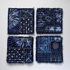May include: Four square fabric coasters featuring deep indigo blue dyed patterns with white hand-stitched embroidery. Each coaster displays a unique patchwork design with frayed edges, incorporating geometric lines, circular motifs, and tie-dye effects.