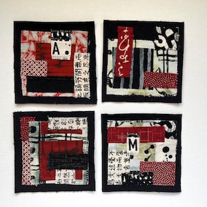 May include: Four square fabric art pieces with black borders. Each features a collage of red, white, and black patterned fabrics, with Japanese text and the letters "A" and "M". The art pieces have a handmade, textured appearance.