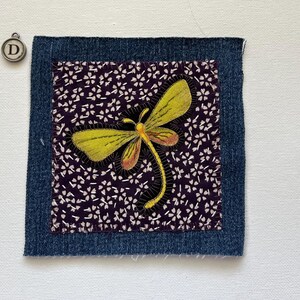 May include: A square fabric patch with a denim border. Inside, a dark purple fabric with white floral patterns surrounds a yellow and orange dragonfly applique. A small metal charm with the letter "D" is in the upper left corner.