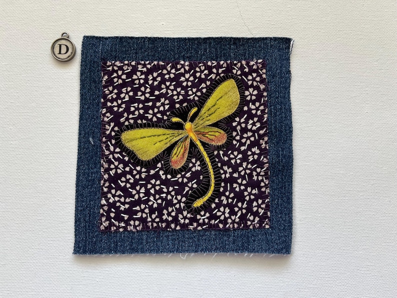 May include: A square fabric patch with a denim border. Inside, a dark purple fabric with white floral patterns surrounds a yellow and orange dragonfly applique. A small metal charm with the letter "D" is in the upper left corner.