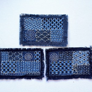 May include: Three rectangular fabric coasters featuring dark blue indigo-dyed cloth with white sashiko-style geometric embroidery patterns. Each piece has a frayed, raw edge finish and displays a patchwork of different stitched designs.