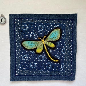 May include: A square, indigo-colored fabric patch with a dragonfly applique. The dragonfly has turquoise wings and a yellow body. The patch is framed with a darker blue denim border and white stitching.