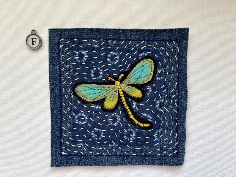 May include: A square, indigo-colored fabric patch with a dragonfly applique. The dragonfly has turquoise wings and a yellow body. The patch is framed with a darker blue denim border and white stitching.