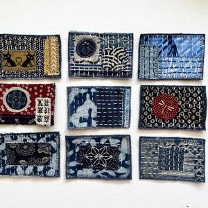 May include: Nine small, rectangular fabric patches with various patterns and textures. The patches feature indigo blue, white, red, and gold colors. Some patches have cat and floral designs, while others have geometric patterns. The patches are hand-stitched with white thread.