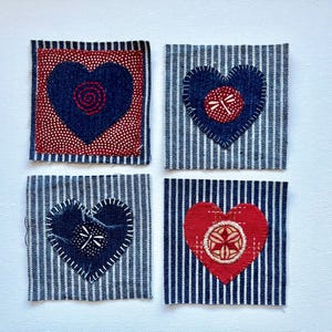 May include: Four square fabric patches with heart designs. Two patches have blue hearts on striped fabric, one with a dragonfly and one with a stitched spiral. The other two have red hearts, one with a white and red design and one with a white design.