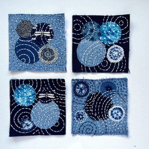 May include: Four square fabric patches with indigo blue and light blue circular designs. Each patch features white stitching and a variety of patterns, including spirals, dots, and dragonfly motifs. The patches are arranged against a white background.