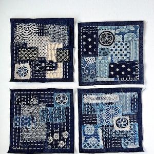 May include: Four indigo blue and white patchwork squares with visible stitching. Each square features a unique combination of patterned fabric pieces, including floral and geometric designs. The squares are arranged against a white background.
