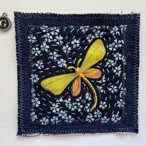 May include: A square fabric patch with a yellow and orange dragonfly design on a dark blue floral background. The patch is framed with dark blue denim and white stitching. A silver charm with the letter "G" hangs nearby.