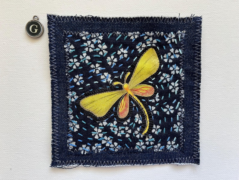 May include: A square fabric patch with a yellow and orange dragonfly design on a dark blue floral background. The patch is framed with dark blue denim and white stitching. A silver charm with the letter "G" hangs nearby.