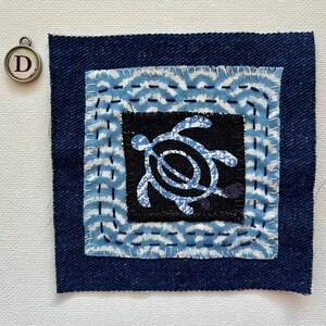 Peut inclure: Pi&egrave;ce d'art textile carr&eacute;e avec une bordure en denim bleu fonc&eacute;. Le centre pr&eacute;sente un carr&eacute; noir avec un motif de tortue bleu clair. La bordure int&eacute;rieure a un motif de vagues en bleu clair et blanc. Un charm argent&eacute; avec la lettre "D" est en haut &agrave; gauche.