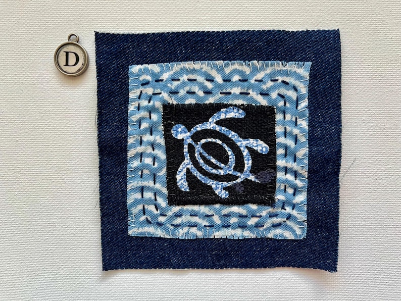 Peut inclure: Pi&egrave;ce d'art textile carr&eacute;e avec une bordure en denim bleu fonc&eacute;. Le centre pr&eacute;sente un carr&eacute; noir avec un motif de tortue bleu clair. La bordure int&eacute;rieure a un motif de vagues en bleu clair et blanc. Un charm argent&eacute; avec la lettre "D" est en haut &agrave; gauche.