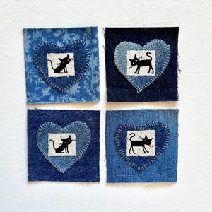 May include: Four denim patches with heart-shaped appliqués. Each heart is stitched onto a square of denim and features a black cat illustration on a white square. The denim varies in shades of blue.