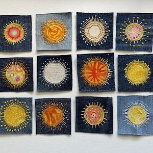 May include: A collection of twelve denim squares, each featuring a unique embroidered sun design. The suns vary in color, including yellow, orange, pink, and white, with detailed stitching. The denim squares are arranged on a white background.