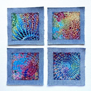 May include: Four square fabric patches featuring a denim border surrounding a colorful center panel. Each center panel displays a vibrant, multi-colored abstract pattern with white thread stitched in a spiral design across the surface.