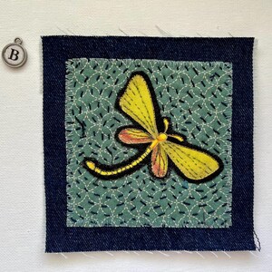 May include: A square fabric patch with a yellow and orange dragonfly design, stitched onto a teal background with a white pattern. The patch is framed by a dark blue denim border. A silver charm with the letter "B" hangs nearby.
